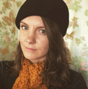 Head shot of a white woman wiht blond hair, a black tam hat, and knitted orange scarf. She stands against butterfly wallpaper.