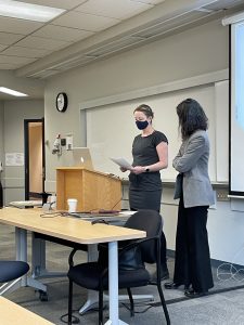 Karis Shearer and Linda Morra, two white women stand at a podium wearing face masks and introducing the event.