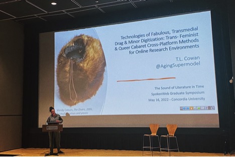 TL Cowan stands at podium on a stage in front of a very large screen showing slide reading "Technologies of Fabulous, Transmedial Drag & Minor Digitization: Trans-Feminist & Queer Cabaret Cross-Platform Methods for Online Research Environments.