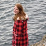 A red-headed woman wearing a red and black plaid jacket looks back over her shoulder at the camera. In front of her is blue lake water.