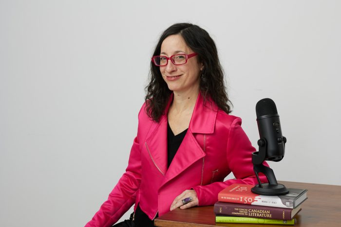 Linda Morra, a white woman with dark brown hair wears a bright pink jacket and sits at a desk with microphone and a stack of three books