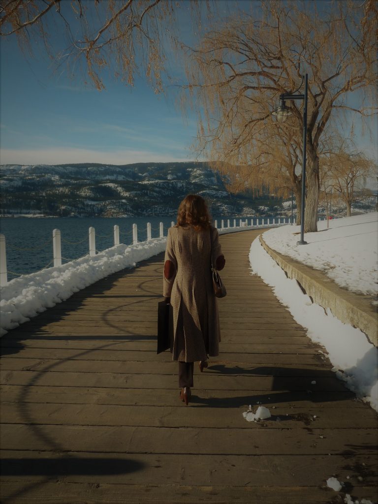 A woman with blond curly hair, wearing long tan coat walks away from the camera on a boardwalk by the lake.