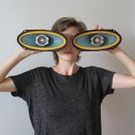 A white woman wearing a black tshirt holds up two large oval shapes over her eyes.