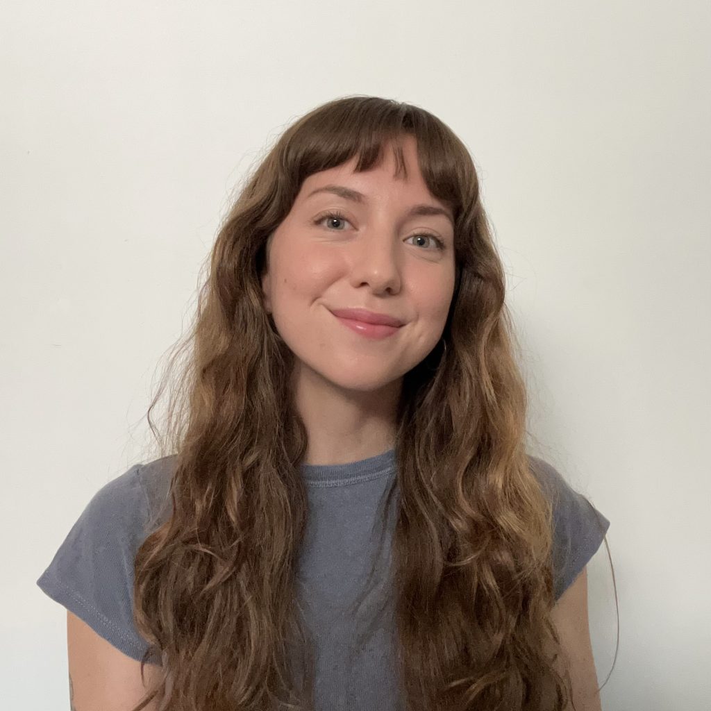 A young white woman with long brown hair and bangs smiles at the camera against a white backgound. She wears a light blue-grey tshirt.
