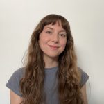 A young white woman with long brown hair and bangs smiles at the camera against a white backgound. She wears a light blue-grey tshirt. 